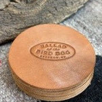 A set of Ballad of the Bird Dog 5 Pack Leather Coasters, each handmade and round with “Ballad of the Bird Dog, Brenham, TX” embossed on them, displayed on a textured surface beside a piece of wood.