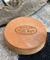 A set of Ballad of the Bird Dog 5 Pack Leather Coasters, each handmade and round with “Ballad of the Bird Dog, Brenham, TX” embossed on them, displayed on a textured surface beside a piece of wood.