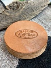 A set of Ballad of the Bird Dog 5 Pack Leather Coasters, each handmade and round with “Ballad of the Bird Dog, Brenham, TX” embossed on them, displayed on a textured surface beside a piece of wood.