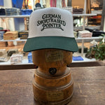 A Ballad Originals Bird Dog Series Hat | German Shorthaired Pointer—featuring white and green colors with “German Shorthaired Pointer” text—is displayed on a wooden hat stand in a store among folded clothes, showcasing Texas craftsmanship inspired by loyal hunting companions.