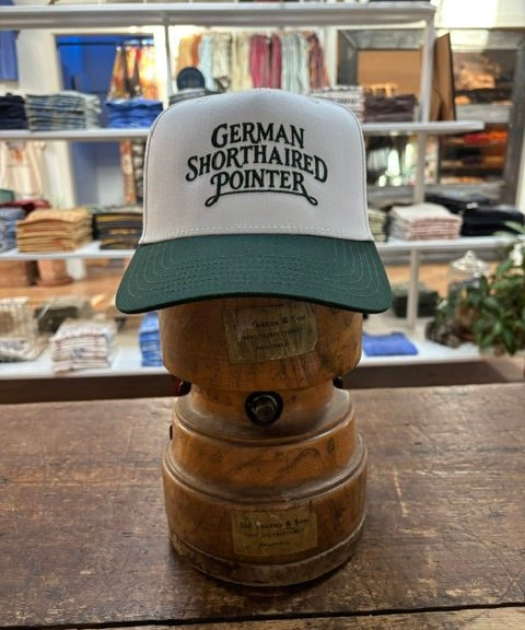 A Ballad Originals Bird Dog Series Hat | German Shorthaired Pointer—featuring white and green colors with “German Shorthaired Pointer” text—is displayed on a wooden hat stand in a store among folded clothes, showcasing Texas craftsmanship inspired by loyal hunting companions.