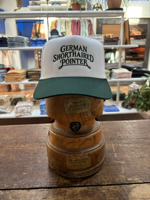 A Ballad Originals Bird Dog Series Hat | German Shorthaired Pointer—featuring white and green colors with “German Shorthaired Pointer” text—is displayed on a wooden hat stand in a store among folded clothes, showcasing Texas craftsmanship inspired by loyal hunting companions.