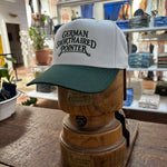 A Ballad Originals Bird Dog Series Hat | German Shorthaired Pointer in white and green is displayed on a wooden hat stand in a store, surrounded by plants and shelves. The hat features “German Shorthaired Pointer” text, celebrating Texas craftsmanship and the loyal hunting companion.