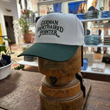 A Ballad Originals Bird Dog Series Hat | German Shorthaired Pointer in white and green is displayed on a wooden hat stand in a store, surrounded by plants and shelves. The hat features “German Shorthaired Pointer” text, celebrating Texas craftsmanship and the loyal hunting companion.