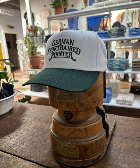 A Ballad Originals Bird Dog Series Hat | German Shorthaired Pointer in white and green is displayed on a wooden hat stand in a store, surrounded by plants and shelves. The hat features “German Shorthaired Pointer” text, celebrating Texas craftsmanship and the loyal hunting companion.