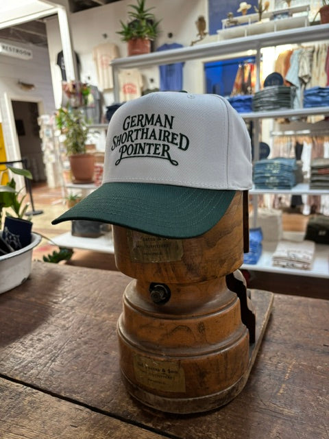 A Ballad Originals Bird Dog Series Hat | German Shorthaired Pointer in white and green is displayed on a wooden hat stand in a store, surrounded by plants and shelves. The hat features “German Shorthaired Pointer” text, celebrating Texas craftsmanship and the loyal hunting companion.