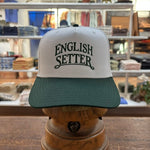 The Bird Dog Series Hat | English Setter by Ballad Originals, featuring a white and green design with “ENGLISH SETTER” printed on the front, is displayed on a wooden stand in a clothing store. Showcasing Texas craftsmanship, this hat is perfect for any hunting companion.