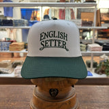 The Bird Dog Series Hat | English Setter by Ballad Originals, featuring a white and green design with “ENGLISH SETTER” printed on the front, is displayed on a wooden stand in a clothing store. Showcasing Texas craftsmanship, this hat is perfect for any hunting companion.