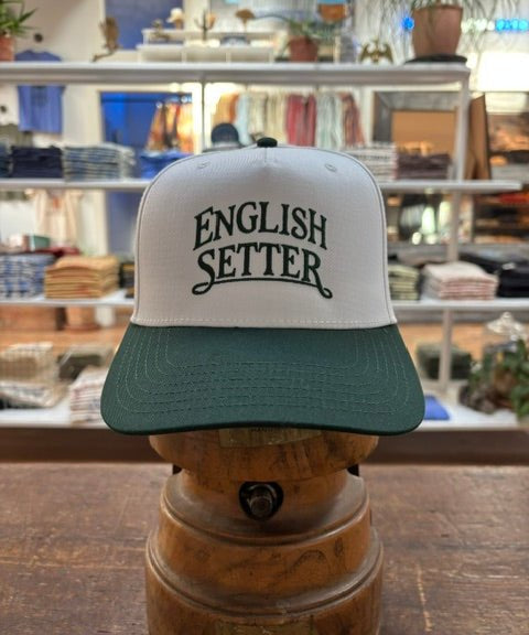 The Bird Dog Series Hat | English Setter by Ballad Originals, featuring a white and green design with “ENGLISH SETTER” printed on the front, is displayed on a wooden stand in a clothing store. Showcasing Texas craftsmanship, this hat is perfect for any hunting companion.