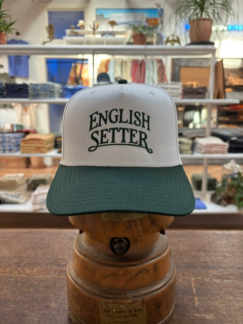 The Bird Dog Series Hat | English Setter by Ballad Originals, featuring a white and green design with “ENGLISH SETTER” printed on the front, is displayed on a wooden stand in a clothing store. Showcasing Texas craftsmanship, this hat is perfect for any hunting companion.