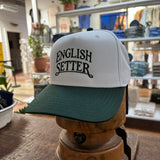 Displayed on a wooden stand inside a retail store, the Ballad Originals Bird Dog Series Hat | English Setter features a white and green design with “ENGLISH SETTER” printed on the front, highlighting Texas craftsmanship.