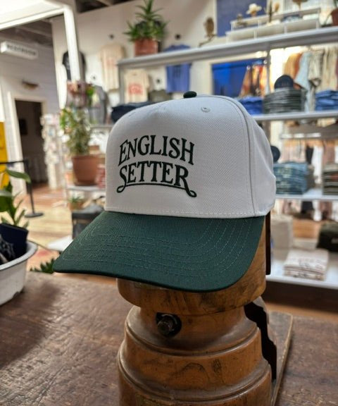 Displayed on a wooden stand inside a retail store, the Ballad Originals Bird Dog Series Hat | English Setter features a white and green design with “ENGLISH SETTER” printed on the front, highlighting Texas craftsmanship.