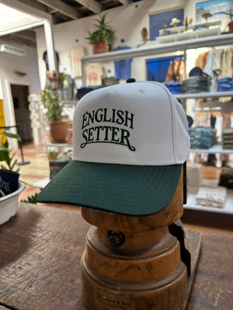 Displayed on a wooden stand inside a retail store, the Ballad Originals Bird Dog Series Hat | English Setter features a white and green design with “ENGLISH SETTER” printed on the front, highlighting Texas craftsmanship.