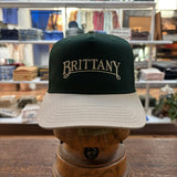 The Ballad Originals Bird Dog Series Hat | Brittany, featuring green and beige tones and “BRITTANY” embroidered on the front to reflect authentic Texas craftsmanship, is displayed on a wooden stand in a clothing store.