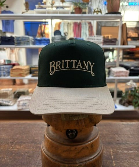 The Ballad Originals Bird Dog Series Hat | Brittany, featuring green and beige tones and “BRITTANY” embroidered on the front to reflect authentic Texas craftsmanship, is displayed on a wooden stand in a clothing store.
