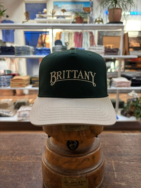 The Ballad Originals Bird Dog Series Hat | Brittany, featuring green and beige tones and “BRITTANY” embroidered on the front to reflect authentic Texas craftsmanship, is displayed on a wooden stand in a clothing store.