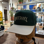 The Bird Dog Series Hat | Brittany by Ballad Originals features a green and beige design with “BRITTANY” embroidered on the front. Displayed on a wooden stand among shelves and plants, this hat highlights authentic Texas craftsmanship.