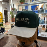 The Bird Dog Series Hat | Brittany by Ballad Originals features a green and beige design with “BRITTANY” embroidered on the front. Displayed on a wooden stand among shelves and plants, this hat highlights authentic Texas craftsmanship.