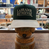 A Bird Dog Series Hat | Labrador Retriever by Ballad Originals, featuring a green and white design with “Labrador Retriever” on the front, is displayed on a wooden hat stand in a clothing store—showcasing classic Texas craftsmanship and making it the perfect signature hat for any hunting companion.