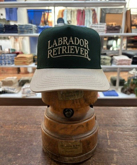 A Bird Dog Series Hat | Labrador Retriever by Ballad Originals, featuring a green and white design with “Labrador Retriever” on the front, is displayed on a wooden hat stand in a clothing store—showcasing classic Texas craftsmanship and making it the perfect signature hat for any hunting companion.