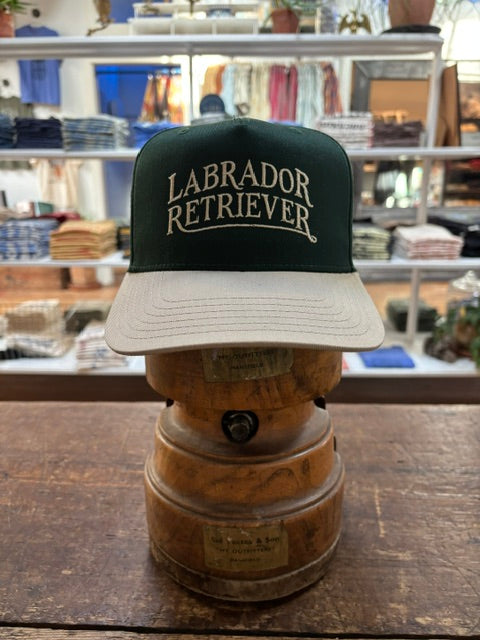 A Bird Dog Series Hat | Labrador Retriever by Ballad Originals, featuring a green and white design with “Labrador Retriever” on the front, is displayed on a wooden hat stand in a clothing store—showcasing classic Texas craftsmanship and making it the perfect signature hat for any hunting companion.