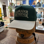 Ballad Originals’ Bird Dog Series Hat | Labrador Retriever—a green and beige cap with “Labrador Retriever” on the front—sits indoors on a wooden stand in the shop, offering a classic tribute to your favorite hunting companion.