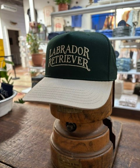 Ballad Originals’ Bird Dog Series Hat | Labrador Retriever—a green and beige cap with “Labrador Retriever” on the front—sits indoors on a wooden stand in the shop, offering a classic tribute to your favorite hunting companion.