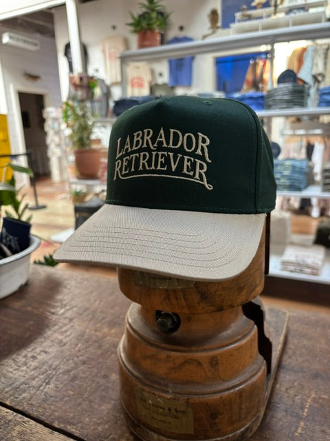 Ballad Originals’ Bird Dog Series Hat | Labrador Retriever—a green and beige cap with “Labrador Retriever” on the front—sits indoors on a wooden stand in the shop, offering a classic tribute to your favorite hunting companion.