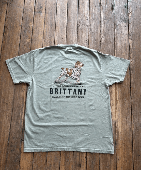 Light gray Bird Dog Series Shirt | Brittany by Ballad Originals on wooden floor, featuring an illustration of a Brittany dog with the text: “True Companionship. BRITTANY. Ballad of the Bird Dog.” Ideal for those who value loyal hunting companions, this tee honors the unique bond between owner and pup.