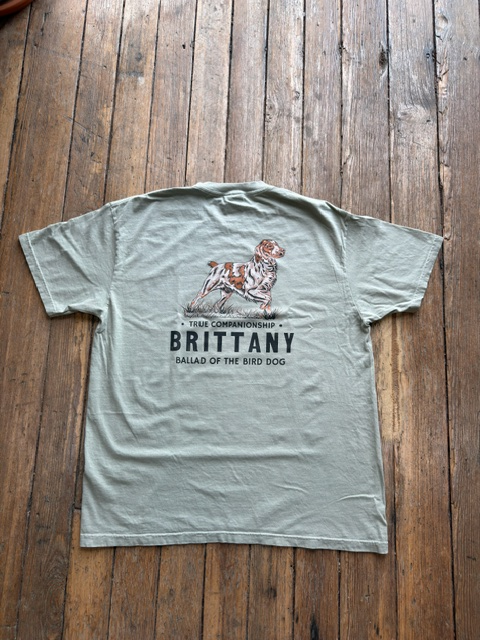 Light gray Bird Dog Series Shirt | Brittany by Ballad Originals on wooden floor, featuring an illustration of a Brittany dog with the text: “True Companionship. BRITTANY. Ballad of the Bird Dog.” Ideal for those who value loyal hunting companions, this tee honors the unique bond between owner and pup.