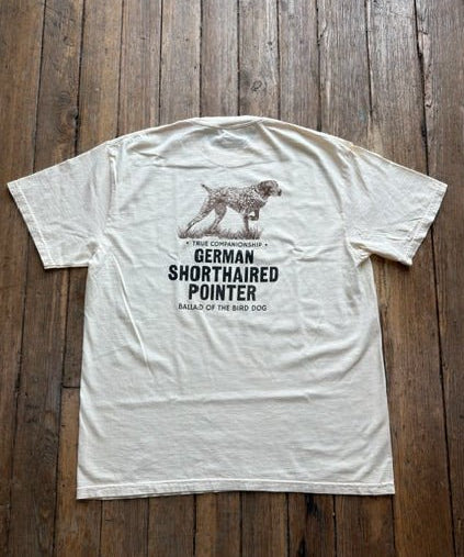 A cream-colored Bird Dog Series Shirt | German Shorthaired Pointer by Ballad Originals lies flat on a wooden floor, featuring a graphic of a dog and the text: “True Companionship, German Shorthaired Pointer, Ballad of the Bird Dog.” Perfect for any bird dog enthusiast, this tee is an ideal hunting shirt or everyday favorite.