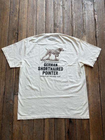 A cream-colored Bird Dog Series Shirt | German Shorthaired Pointer by Ballad Originals lies flat on a wooden floor, featuring a graphic of a dog and the text: “True Companionship, German Shorthaired Pointer, Ballad of the Bird Dog.” Perfect for any bird dog enthusiast, this tee is an ideal hunting shirt or everyday favorite.