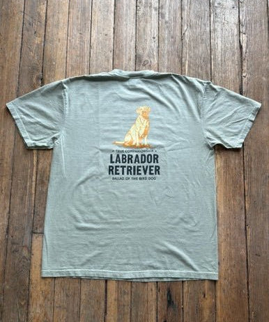 Showcased on a wooden floor, the Ballad Originals Bird Dog Series Shirt | Labrador Retriever features a soft gray Comfort Colors tee with a yellow Labrador Retriever graphic and “Labrador Retriever, Sailor of the Bird Dog” printed on the back—a perfect tribute to loyal hunting companions.
