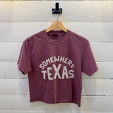 A berry maroon Somewhere Tx Crop Ballad of the Bird Dog cropped t-shirt with “SOMEWHERE TEXAS” in white lettering, hanging on a wooden hanger against a white wood-plank wall.