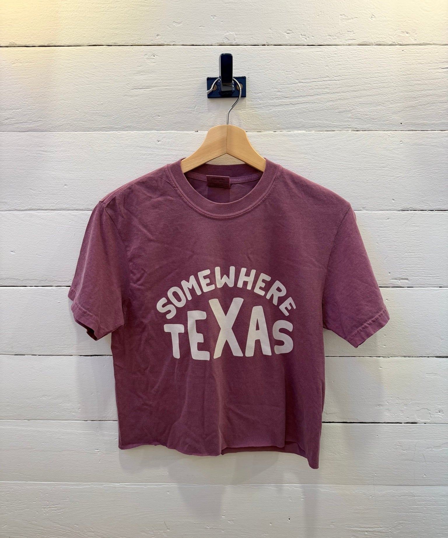 A berry maroon Somewhere Tx Crop Ballad of the Bird Dog cropped t-shirt with “SOMEWHERE TEXAS” in white lettering, hanging on a wooden hanger against a white wood-plank wall.