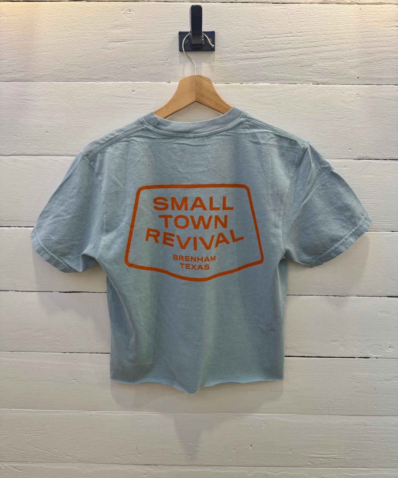 Yankey blue Small Town Revival Crop Ballad of the Bird Dog t-shirt with an orange graphic on the back that reads “Small Town Revival, Brenham Texas” inside a rectangular border.