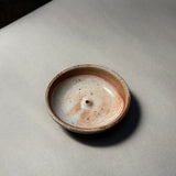 The Incausa Stoneware Holder, a small round incense burner with a beige and brown speckled glaze, sits on a light gray surface, viewed from above.