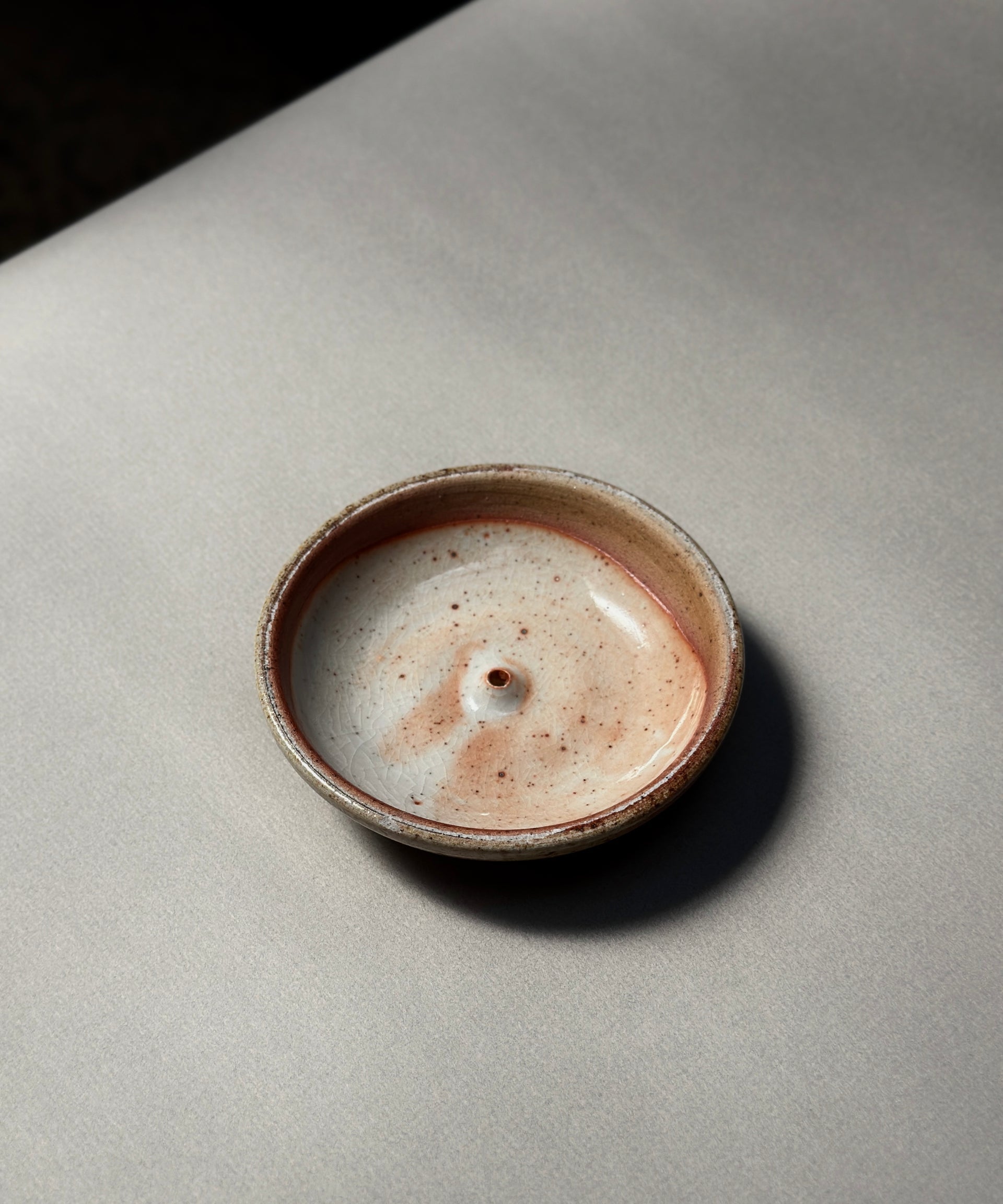 The Incausa Stoneware Holder, a small round incense burner with a beige and brown speckled glaze, sits on a light gray surface, viewed from above.