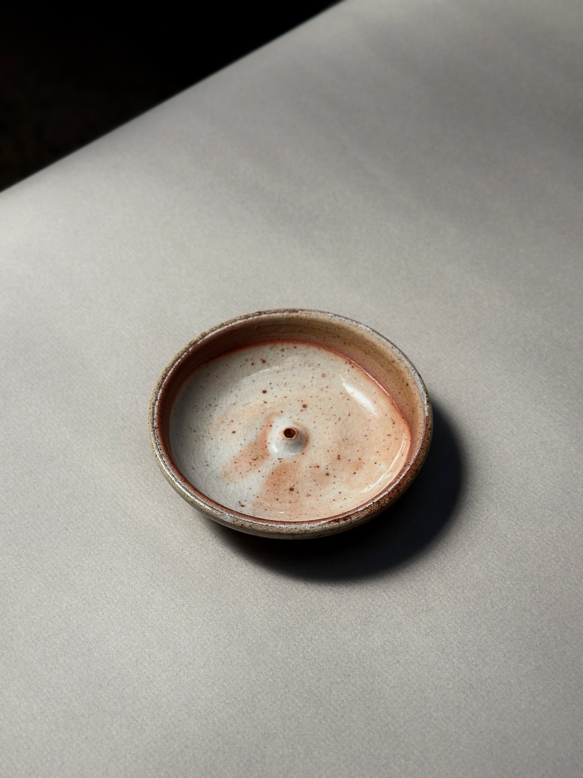 The Incausa Stoneware Holder, a small round incense burner with a beige and brown speckled glaze, sits on a light gray surface, viewed from above.