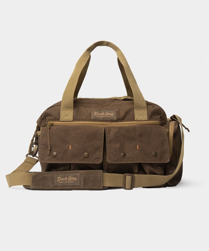 Canvas Blind Bag by Duck Camp in brown waxed canvas, featuring two front pockets, carrying handles, an adjustable shoulder strap, and the “Duck Camp” brand name embroidered on the bag.