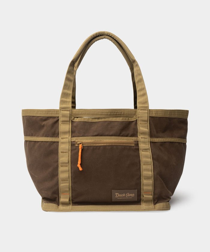 The Canvas Gear Tote by Duck Camp features a brown and tan waxed canvas construction, dual handles, a front zippered pocket with an orange pull, and a “Duck Camp” logo patch at the bottom—making it the ideal durable gear bag for everyday adventures.