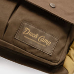 Close-up of the Duck Camp Canvas Upland Strap Vest—a brown bag with a front pocket and a Duck Camp label reading “Premium Outdoor Goods” in yellow lettering—highlighting the rugged durability characteristic of quality field gear.