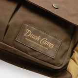 Close-up of the Duck Camp Canvas Upland Strap Vest—a brown bag with a front pocket and a Duck Camp label reading “Premium Outdoor Goods” in yellow lettering—highlighting the rugged durability characteristic of quality field gear.