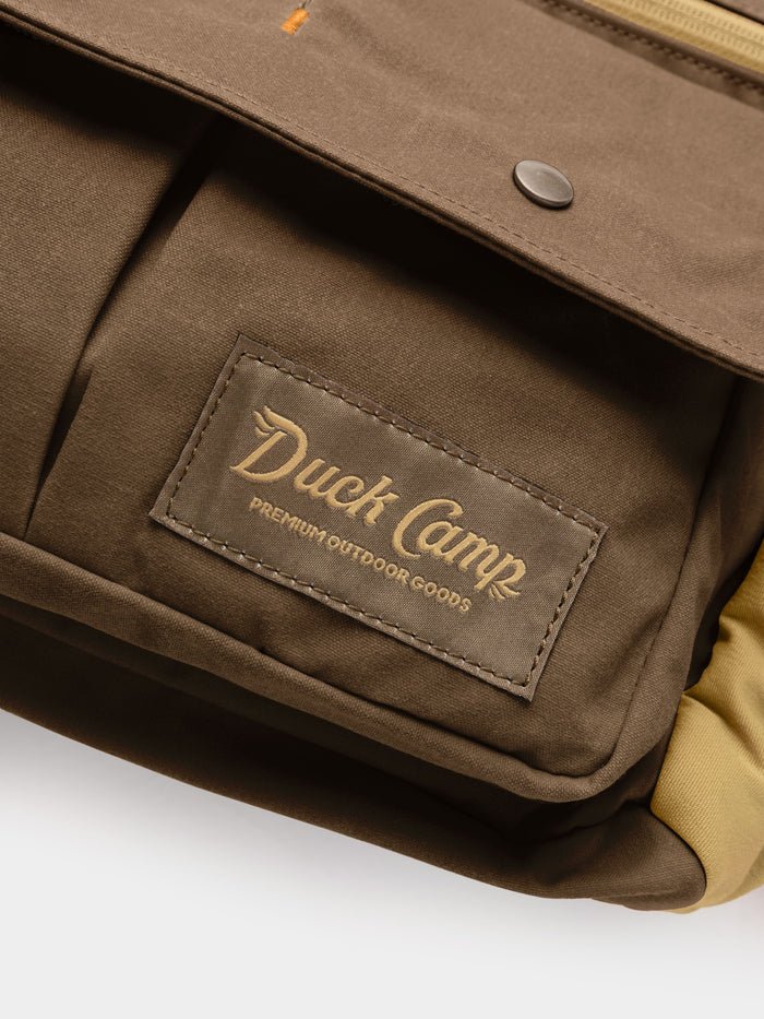 Close-up of the Duck Camp Canvas Upland Strap Vest—a brown bag with a front pocket and a Duck Camp label reading “Premium Outdoor Goods” in yellow lettering—highlighting the rugged durability characteristic of quality field gear.