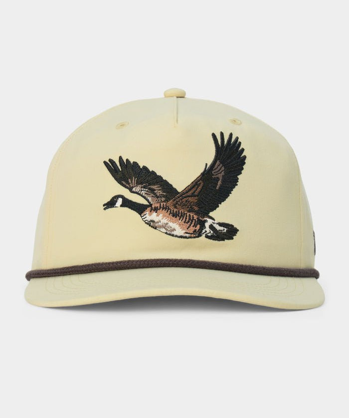 The Canada Goose Hat by Duck Camp is a light yellow, classic 5-panel baseball cap featuring an embroidered flying goose on the front and a dark brown cord above the brim, complete with an adjustable snapback for a perfect fit.