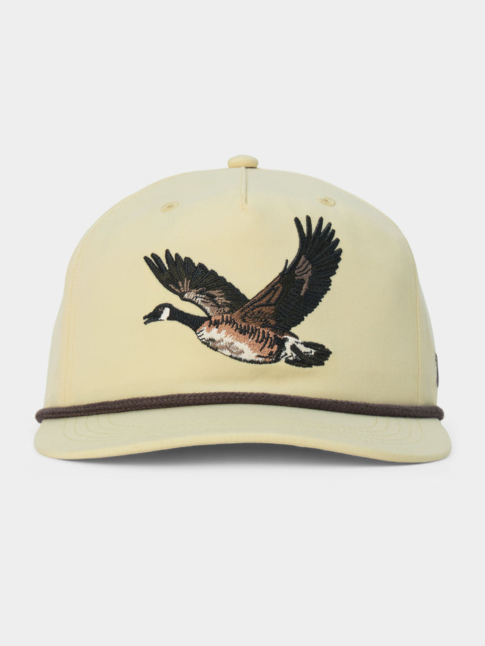 The Canada Goose Hat by Duck Camp is a light yellow, classic 5-panel baseball cap featuring an embroidered flying goose on the front and a dark brown cord above the brim, complete with an adjustable snapback for a perfect fit.