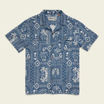 The Shores Club Shirt by Howler Brothers is a short-sleeve blue camp collar shirt with white tropical prints, featuring palm trees and geometric patterns. Made from 100% cotton with a vintage textured weave, shown laid flat on a plain background.