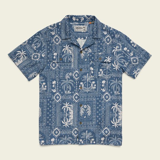 The Shores Club Shirt by Howler Brothers is a short-sleeve blue camp collar shirt with white tropical prints, featuring palm trees and geometric patterns. Made from 100% cotton with a vintage textured weave, shown laid flat on a plain background.