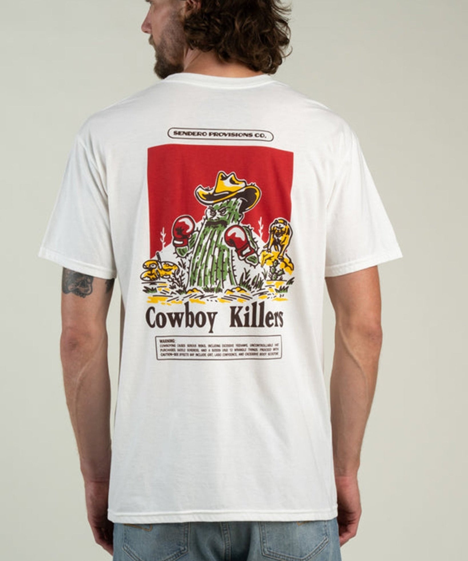 A man wearing the Cowboy Killers T-Shirt by Sendero Provisions Co.—a white organic cotton tee featuring a cartoon cactus with boxing gloves and “Cowboy Killers” printed on the back, finished with a soft-touch screen print for lasting comfort.