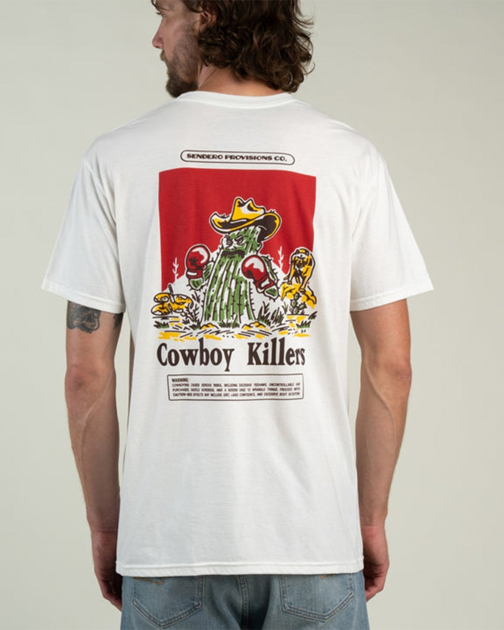 A man wearing the Cowboy Killers T-Shirt by Sendero Provisions Co.—a white organic cotton tee featuring a cartoon cactus with boxing gloves and “Cowboy Killers” printed on the back, finished with a soft-touch screen print for lasting comfort.